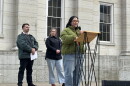 Woman in green jacket talking into mic at outside podium. Two people behind her.
