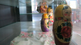 A Russian doll sits on a glass shelf in a glass display case with two smaller dolls behind it 