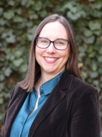 a white woman with dark hair and glasses wearing a teal colored shirt and brown velvet coat smiles and stands in front of a dark ivy background