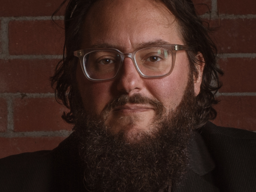 A man with shoulder-length hair and glasses poses for a portrait against a brick wall.