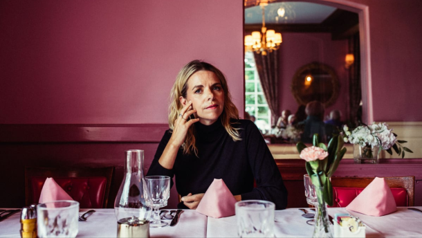 Aoife O'Donovan sits at a dining table in a pink room, looking at the camera with one hand on her cheek.
