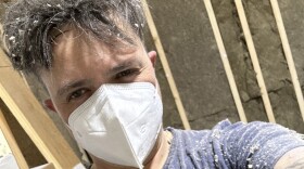 Person covered in sawdust wearing a ventilation mask looking into the came with bemused expression