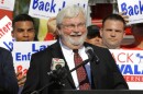 cherub looking older man with white beard and hair wearing wire glasses in dark suit speaks at a podium . behind him are several people holding signs such as "Back Jack" and "Jack Latvala for Governor" 