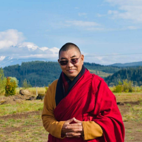 A buddhist monk smiles at the camera with his fingers interlocked with mountains in the background