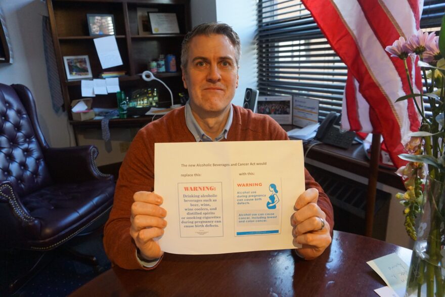 Rep. Andrew Gray, D-Anchorage, in his Capitol office on May 16, 2024, holds up a printout showing how warning signs about alcohol consumption's health effects would change under legislation he sponsored in last year's session.