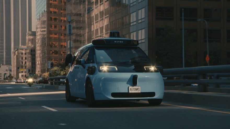 A squat, compact van with an array of autonomous vehicle censors drives down a city street. 