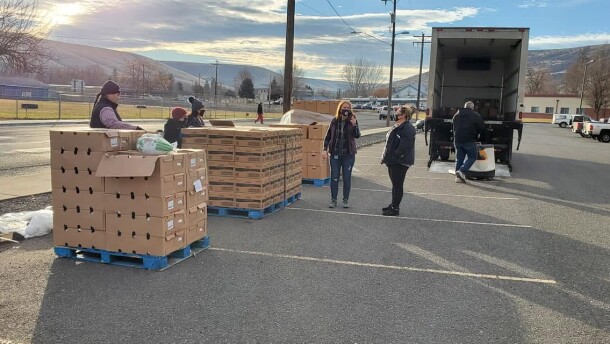 Volunteers stand in a parking lot next to stacks of boxes on pallets. Some boxes are open and an empty truck is nearby. 
