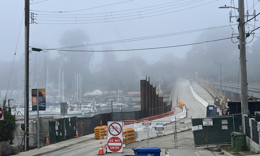 Fencing blocks off the right lane of a bridge while the left side is open. Sailboats float in water on the left side of the image.
