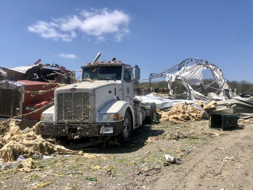 In the foreground, two semi trucks covered with dust and tree branches at the city's tornado-damaged recycling facility. Debris of what remains of the building is in the background.