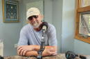 A bearded man in a blue shirt, sunglasses and baseball cap looks into the camera. He is sitting behind a microphone in a radio studio.
