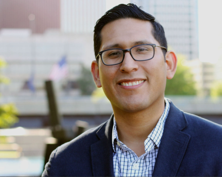 Tony Vargas is a Latino man wearing glasses, a checked shirt and dark glasses. He is smiling in front of a sunny background of tall buildings.