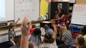 Teacher in front of students gathered on floor. Elementary students raise their hands.