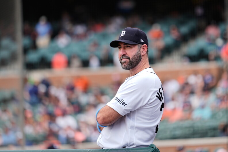 Detroit Tigers pitcher Justin Verlander in Detroit Tigers baseball cap and uniform looks to the right, arms crossed, with fans in the background.