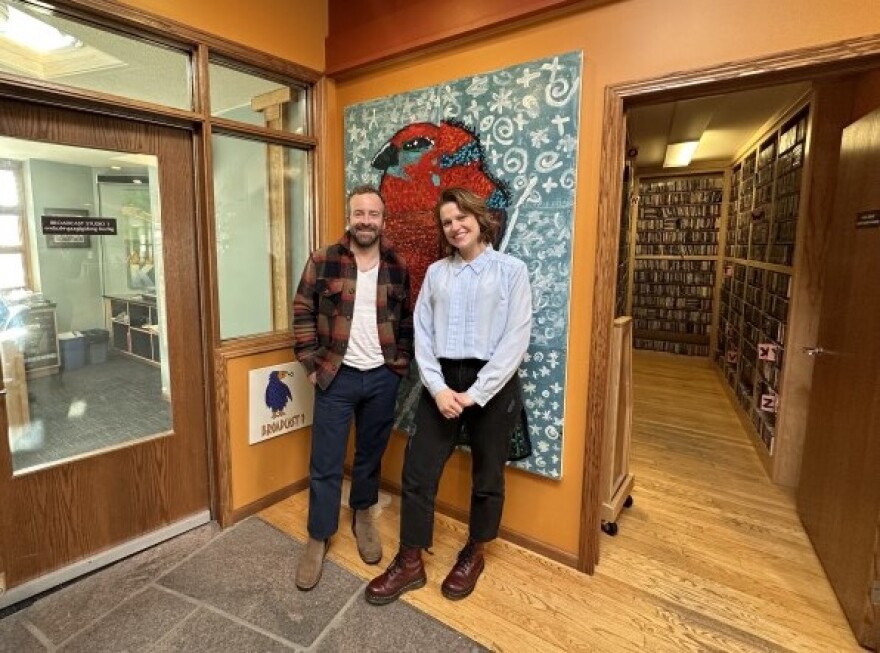 Dave Simonett (wearing dark blue pants and a white V-neck t-shirt under a brown flannel) and Kari Hedlund (wearing dark pants and a light blue button up) smile in front of a painting of a red bird. 