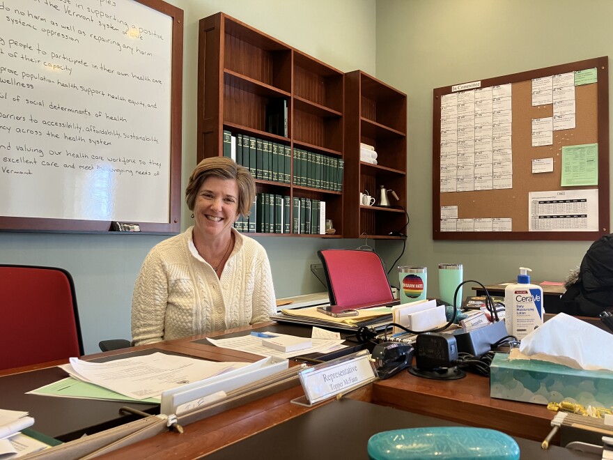 A smiling woman sits at a large cluttered table in a committee room at the Vermont Statehouse.