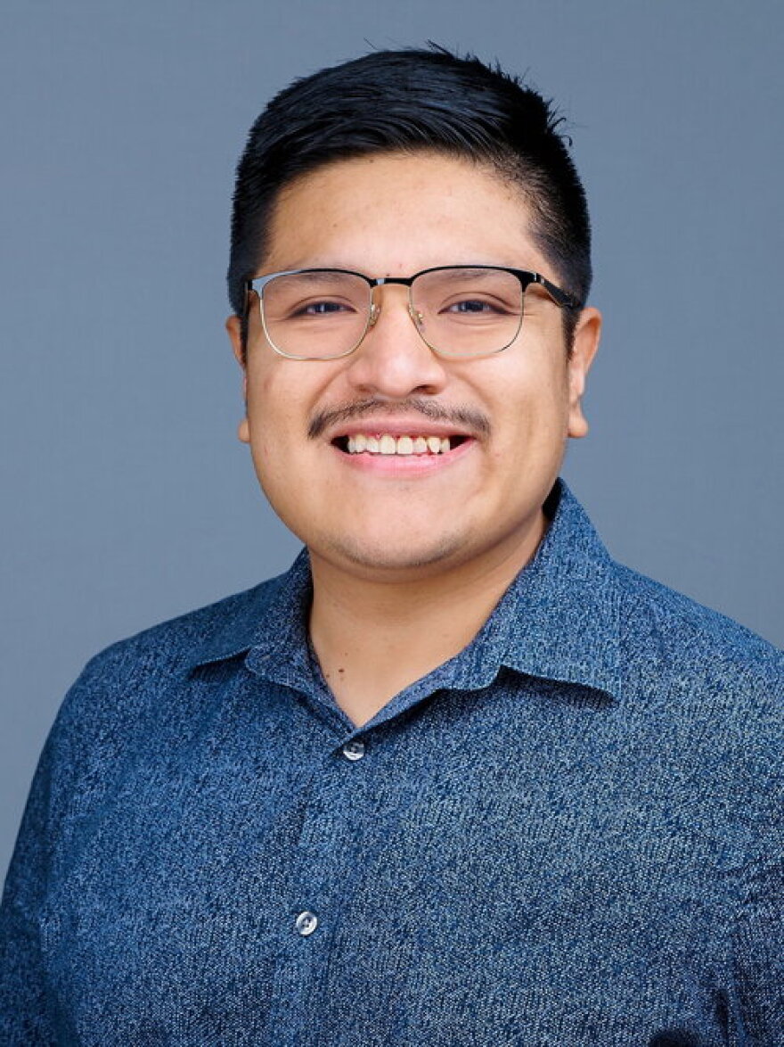 a man with a mustache and glasses wearing a blue polo against a blue-grey background