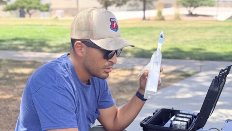 Juan Hernandez, a man wearing a blue T-shirt, green pants, and a tan ball cap, sits in front of a laptop computer outside. He is an incident meteorologist with the National Weather Service, uses a radiosonde while working near the Aspen Acres Fire in southern Colorado on Tuesday. A radiosonde measures temperature, relative humidity, wind and pressure.