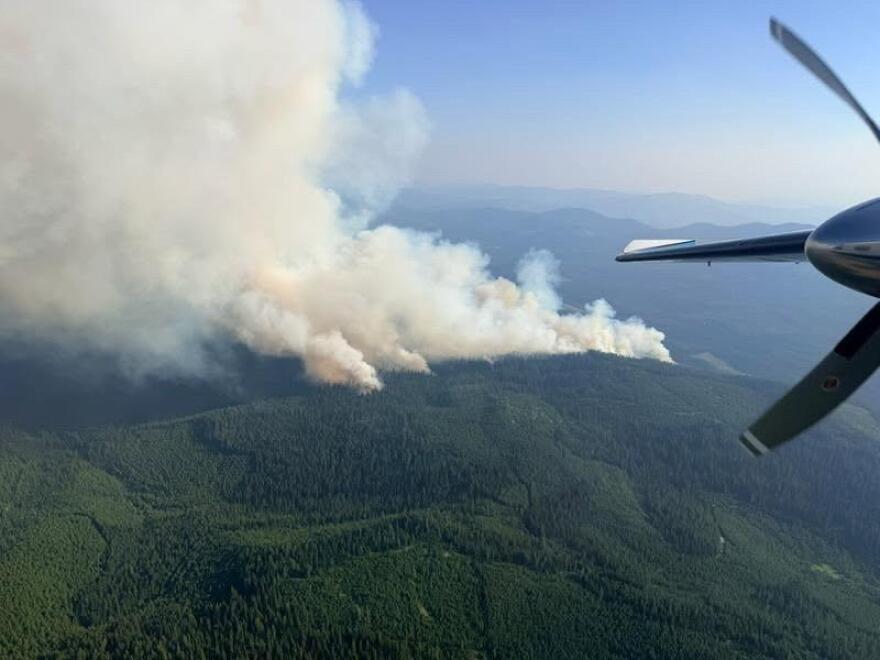 Aerial view from a fire spotting plane. A large plume of smoke rises from green, tree-covered mountains. Part of an airplane propeller is visible in the frame. 