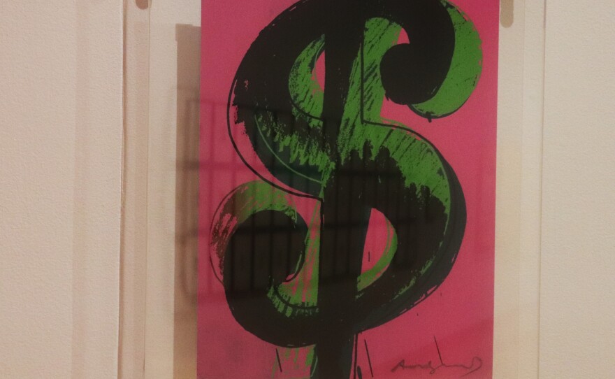 A painting of a green dollar sign with a pink background hangs on a white wall. A row of black-and-white photos are visible in the frame's reflection. 