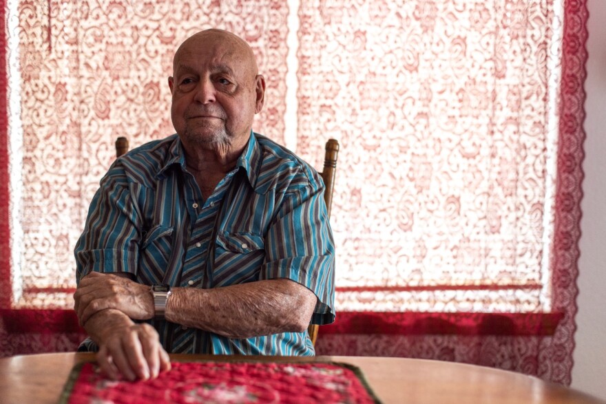 An older man in a striped shirt is seated in front of a floral red window covering. 