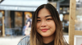 A brown-haired Filipino woman with a black shirt and jean jacket smiling in front of a restaurant