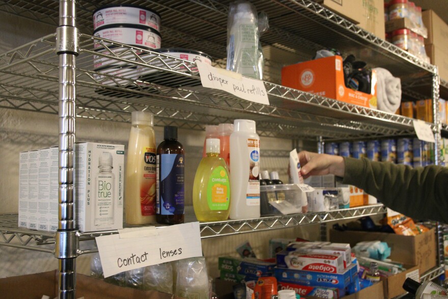 A metal shelf has several toiletry bottles on it, like shampoo and contact cleaner fluid. There's a paper sign that reads "contact lenses." A hand is putting an item on the shelf.