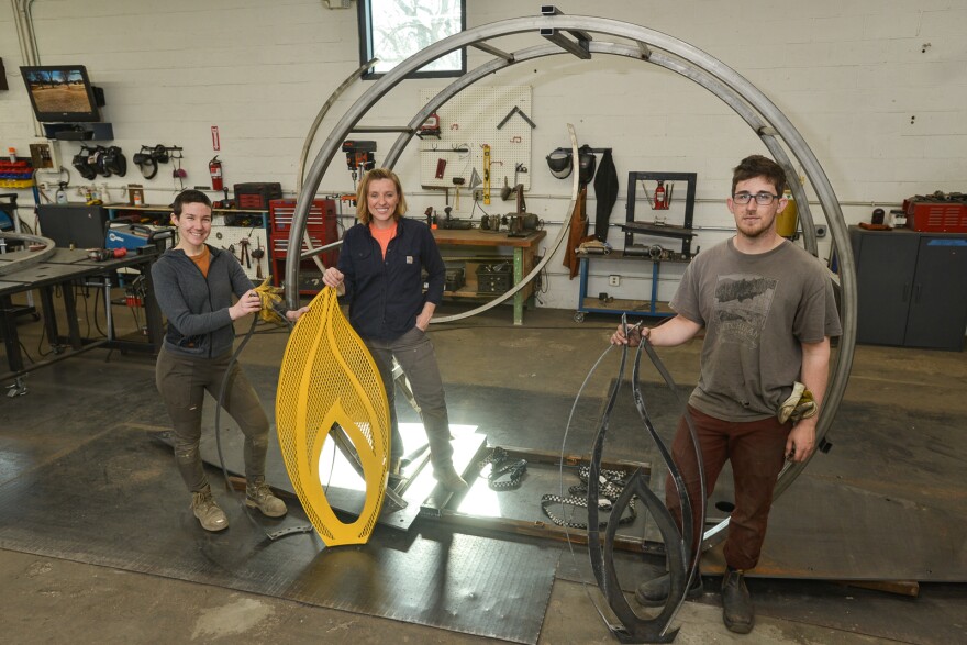 Three people pose with different steel parts that are shaped like sunflower petals. Behind them is a large steel ring and tools and equipment used for metalwork.