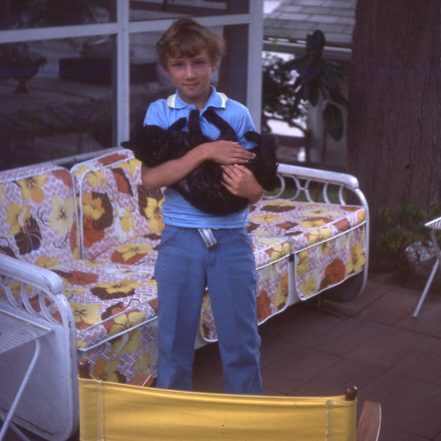 Small boy holding a black cat. Behind him is a rattan couch with floral-printed cushions.