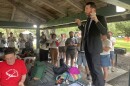 A man stands on a bench to address people under a pavilion
