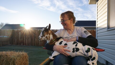 A woman holds a miniature goat dressed in a white onesie with hearts on it.