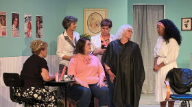 Six women gather in a 1980s beauty salon with fashion posters hung on turquoise walls. Two of the women sit at a manicure table while the others stand around them in conversation.