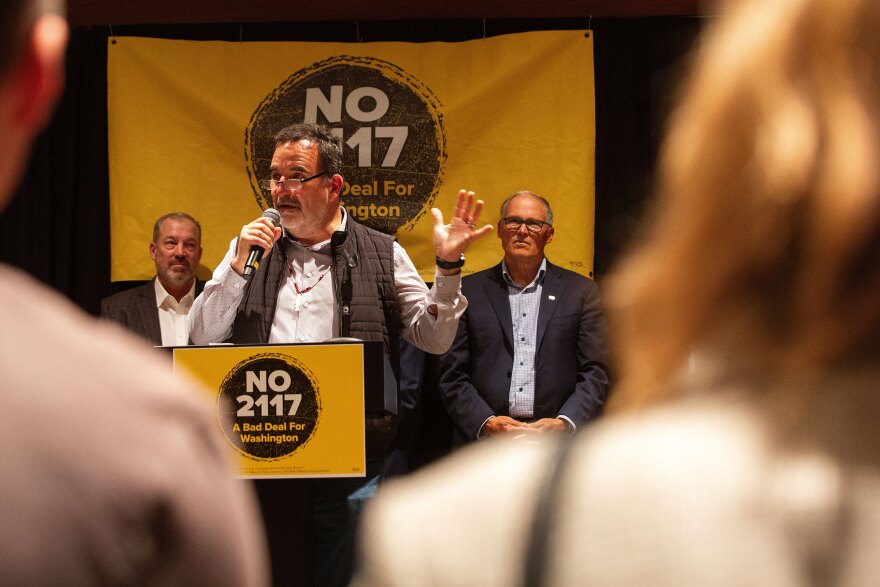 Suquamish Tribal Chairman Leonard Forsman stands at a podium and speaks at the "No on I-2117" campaign party in Seattle on election night, Nov. 5, 2024. Washington Gov. Jay Inslee sits to his right. Yellow and black campaign signs state: No 2117.