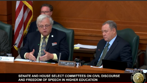Two men suits behind a desk, with another man behind the two