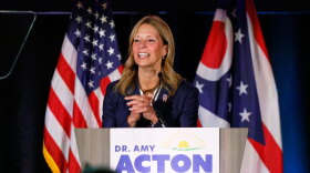 Dr. Amy Acton stands at a podium with a sign reading "Dr. Amy Acton for Governor." She is speaking in front of American and Ohio state flags.