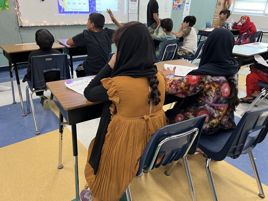 Elementary students sit in rows of two adjoining desks facing a whiteboard. In the foreground are two girls with head scarves. In the background a boy raises his hand.