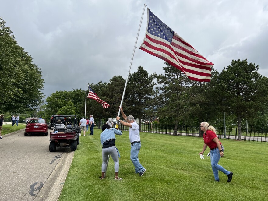 Two people hold up a metal flag pole with an American flag flying above them at the Dayton National Cemetery. Other volunteers are putting up flags in the distance.