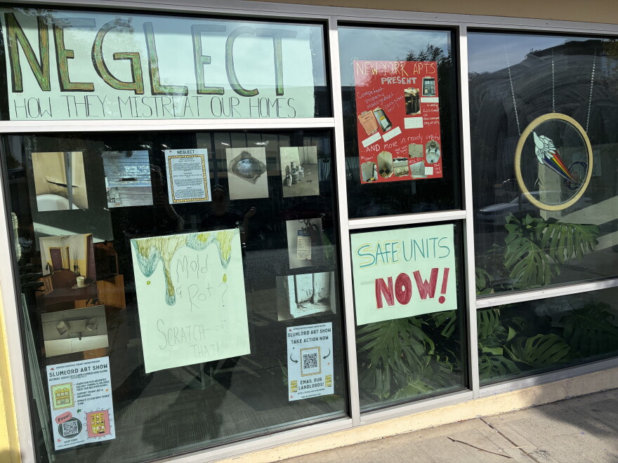 A window that has several signs facing the sidewalk. The signs say"Safe units now!" and "NEGLECT How they mistreat our homes." There are also photos of mold, broken appliances and other substandard conditions. 