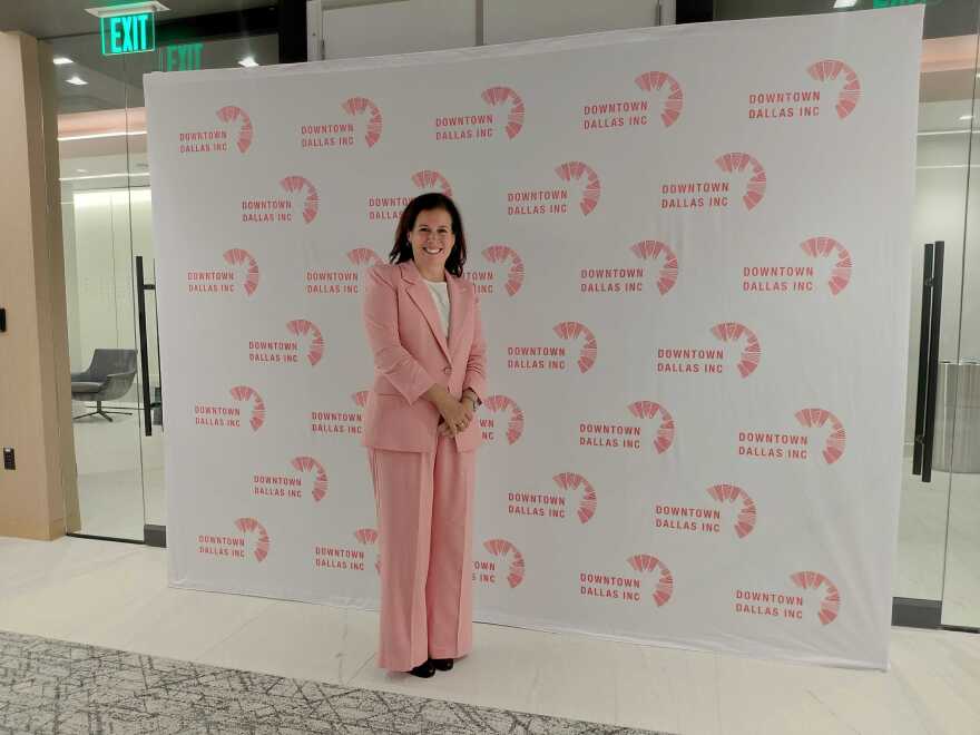 A woman wearing a pink pantsuit stands in front of a backdrop that reads Downtown Dallas, Inc. 