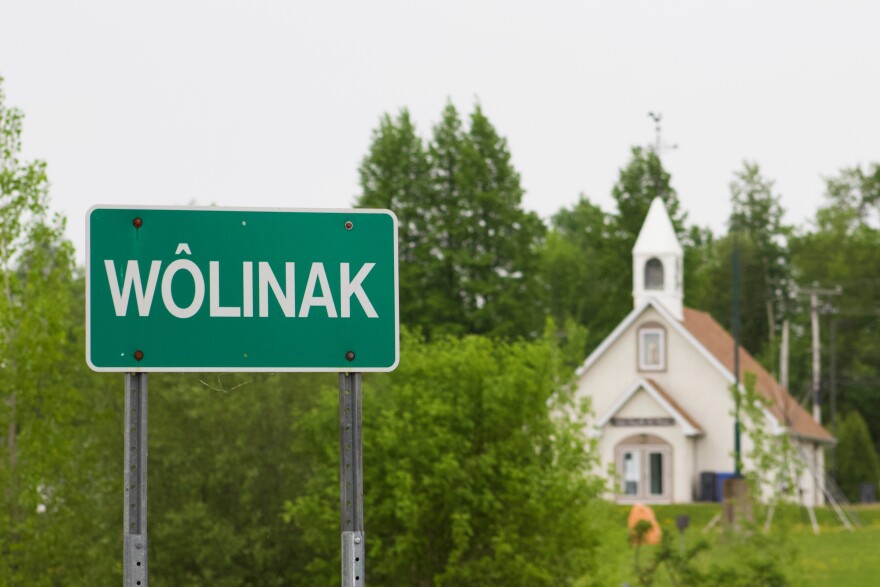 A photo of a green sign with white lettering reading Wôlinak with a white church building and green trees in the background.