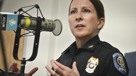 A woman wearing a Kansas City Police uniform sits inside a radio studio. She gesturing with both hands and talking at a microphone.