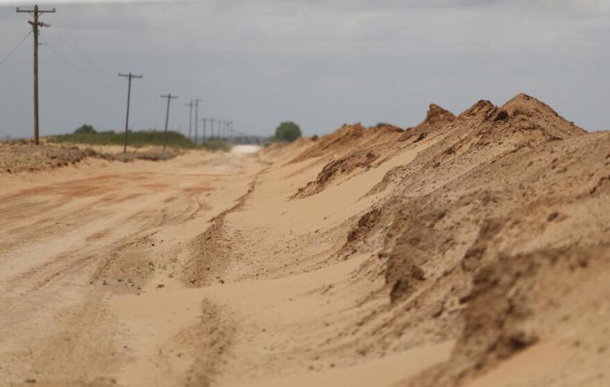 Large piles of sand sit along a country road. Telephone poles line the road opposite the hills of sand.