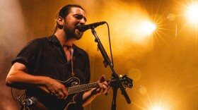 A man plays a mandolin and sings into a microphone on stage with several bright lights in the background.
