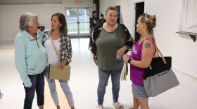 Four white women of varying ages stand together, chatting amiably, in a large linoleum-floored multipurpose room. 