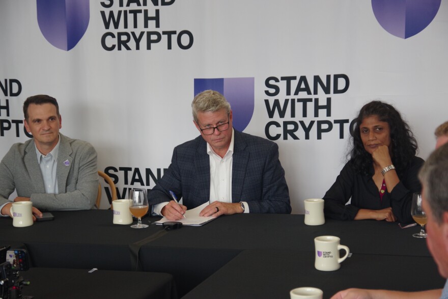 Several people sit around a black table and appear listening to a speaker who is off camera. Behind them, large white banners read "Stand with Crypto"