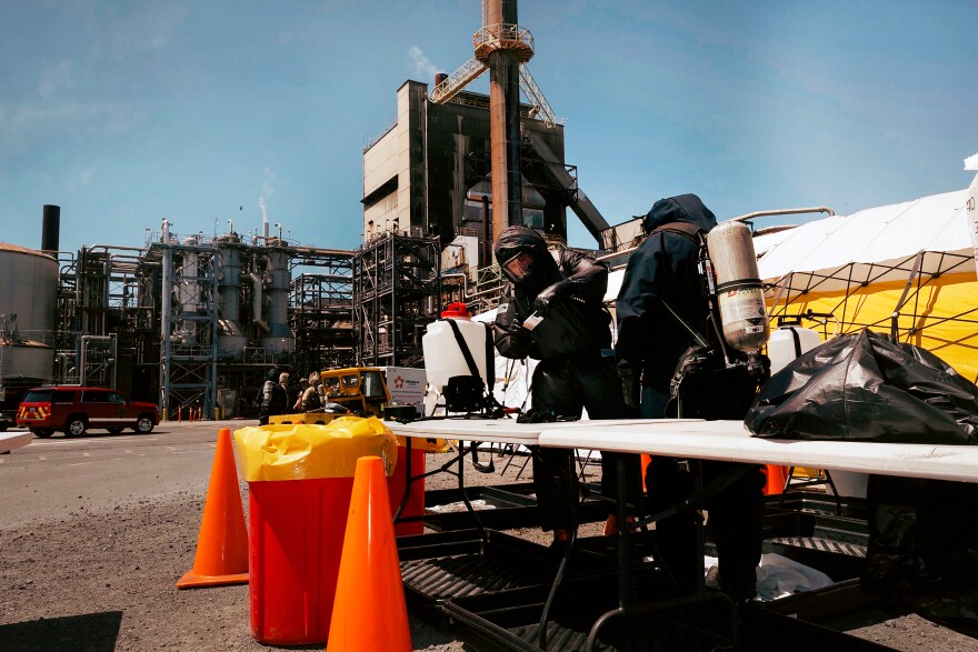 Two people in protective gear with tanks on their back stand with an industrial site in the distance.