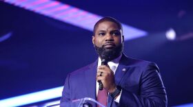 African American man in blue suit holds microphone at podium 