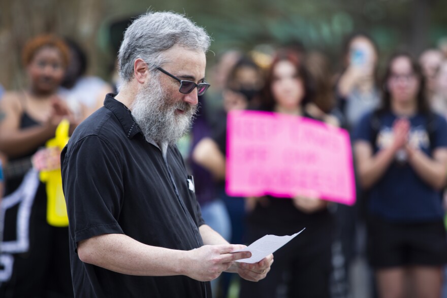 A man with gray hair and a gray beard and glasses reads from a piece of paper to a crowd. 