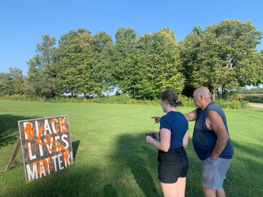 A woman and a man stand looking at a vandalized Black Lives Matter sign.