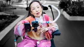 a woman in brightly colored, loose-fitting clothes, with a half-smile and brightly colored lipstick, sitting in a wheelchair with a breathing tube going from her neck to the back of the chair