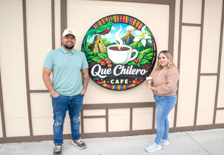 Two people stand outside next to a large Que Chilero logo.